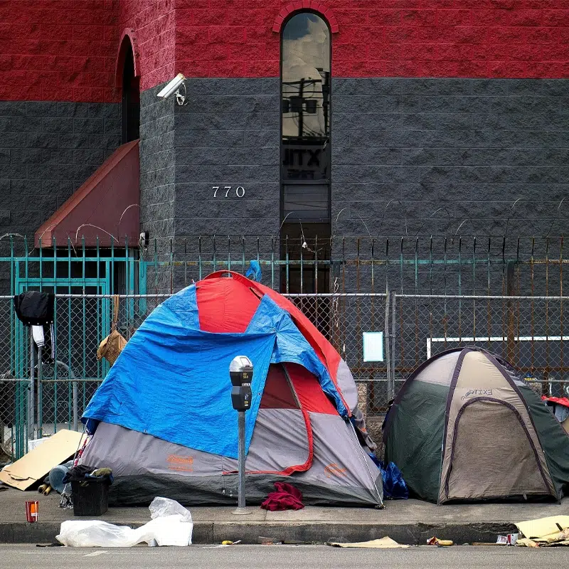 Small tents on an urban street. There is rubbish around the tents and a CCTV camera can be seen above.