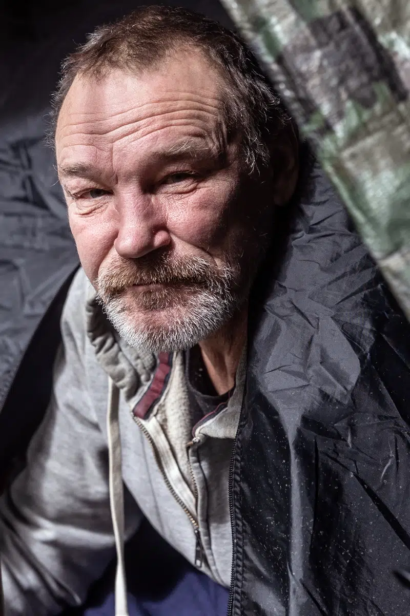 A man in the opening of a make shift tent in a wooded area.
