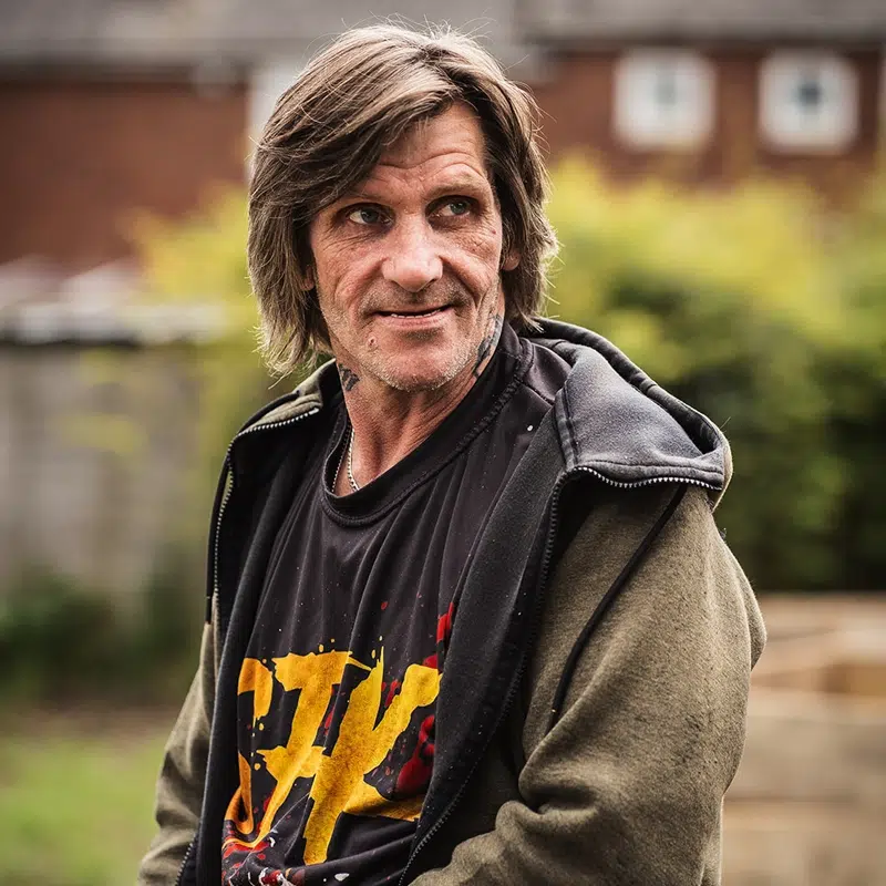 A middle aged man wearing a hoodie sitting outside with houses in the background