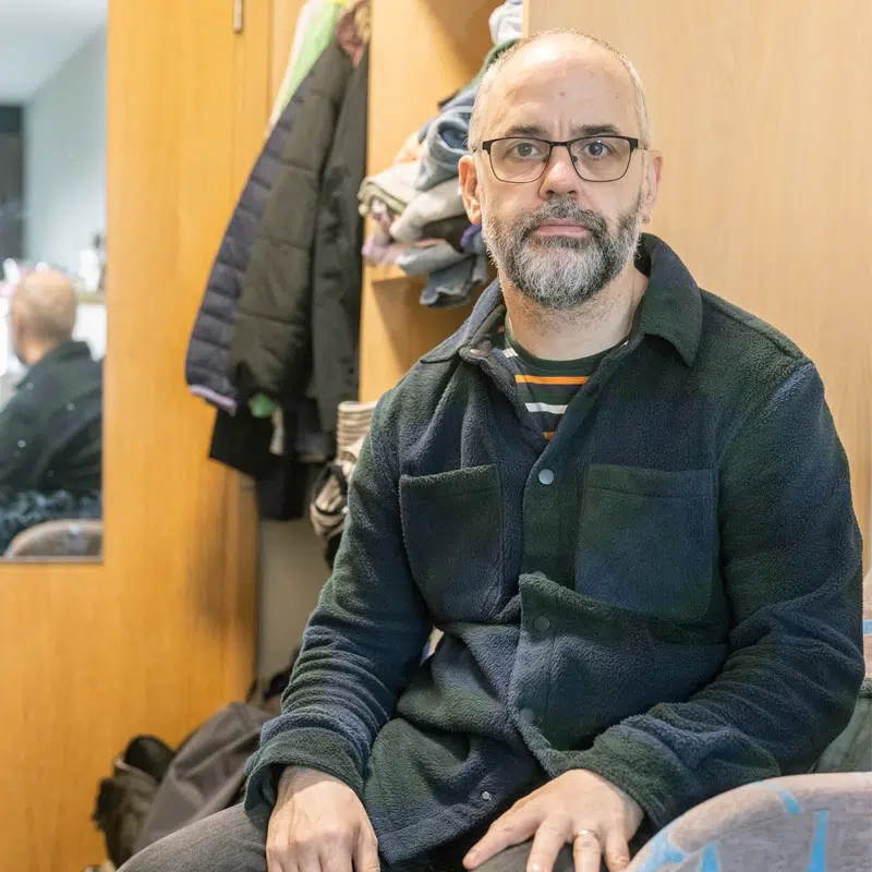 A man sits in a hotel room. Behind him the wardrobe door is open showing his clothes.