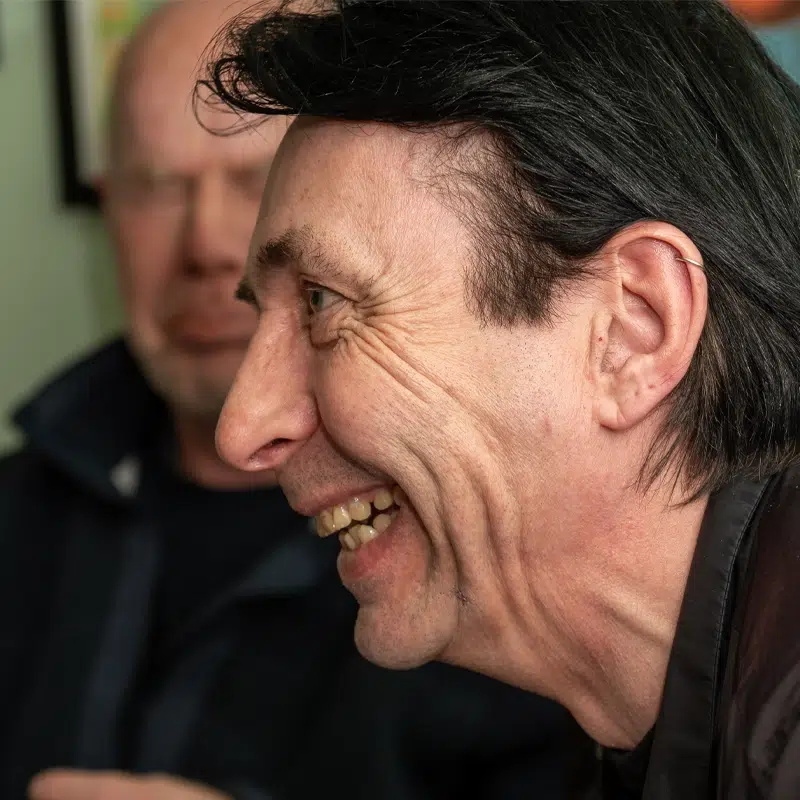 The profile of a male worker smiling. In the background another man's face can be partially seen.