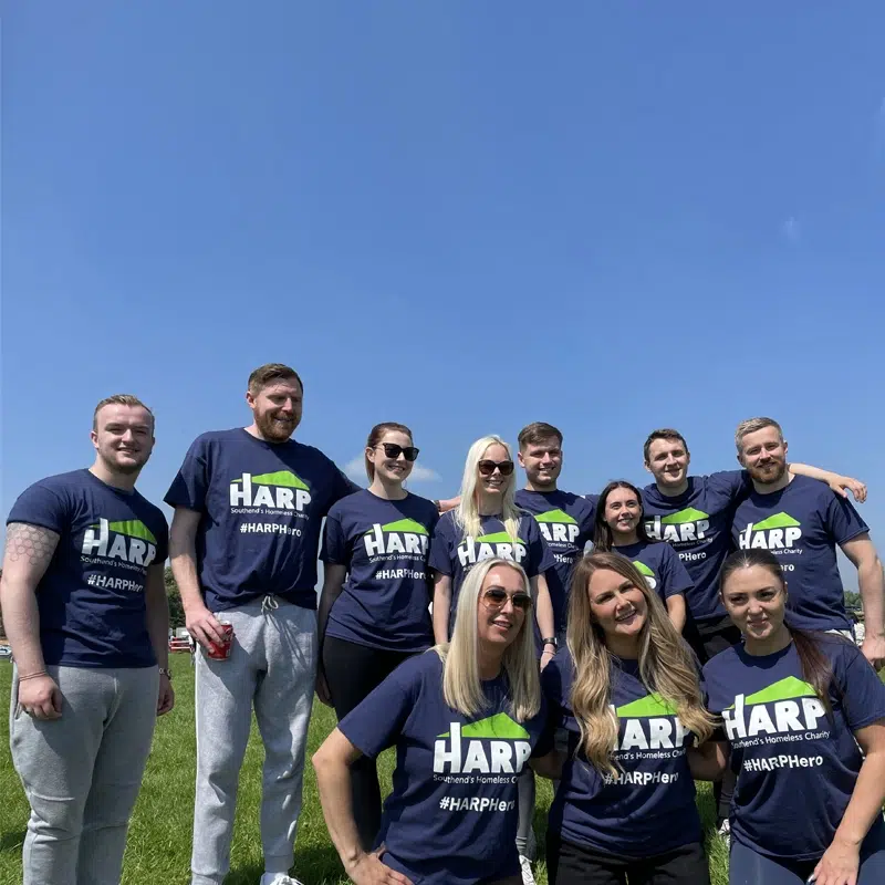 A team of eleven men and women wearing HARP Hero tee-shirts pose as a group in a field against blue skies
