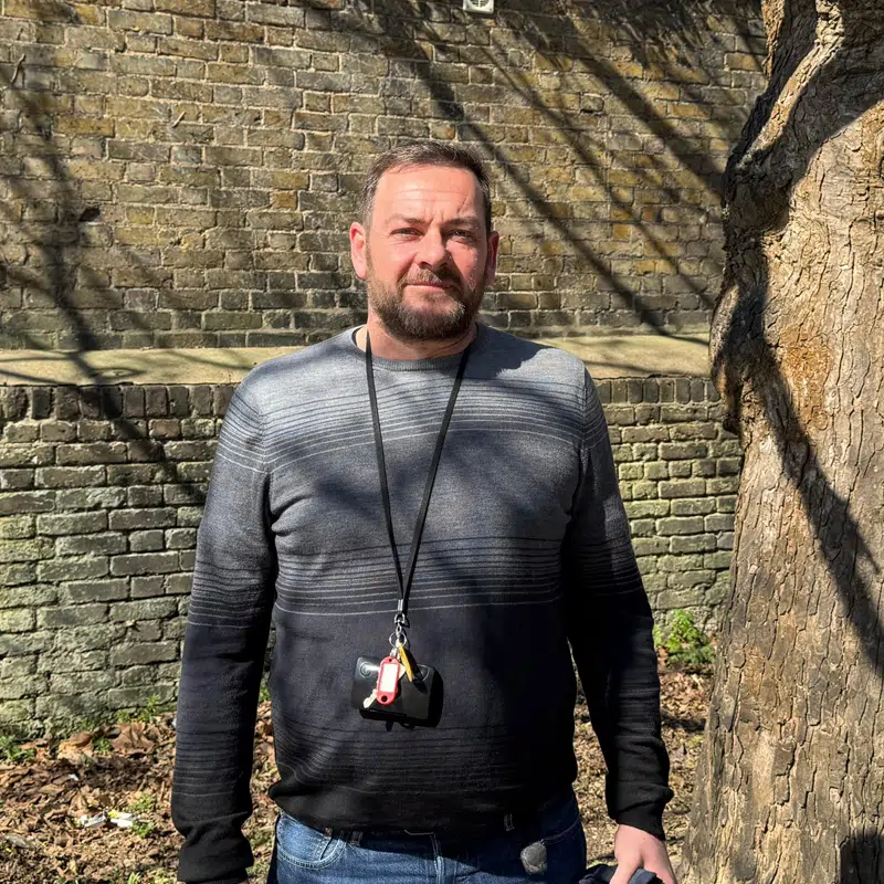 A man standing outside next to a tree in front of a brick wall