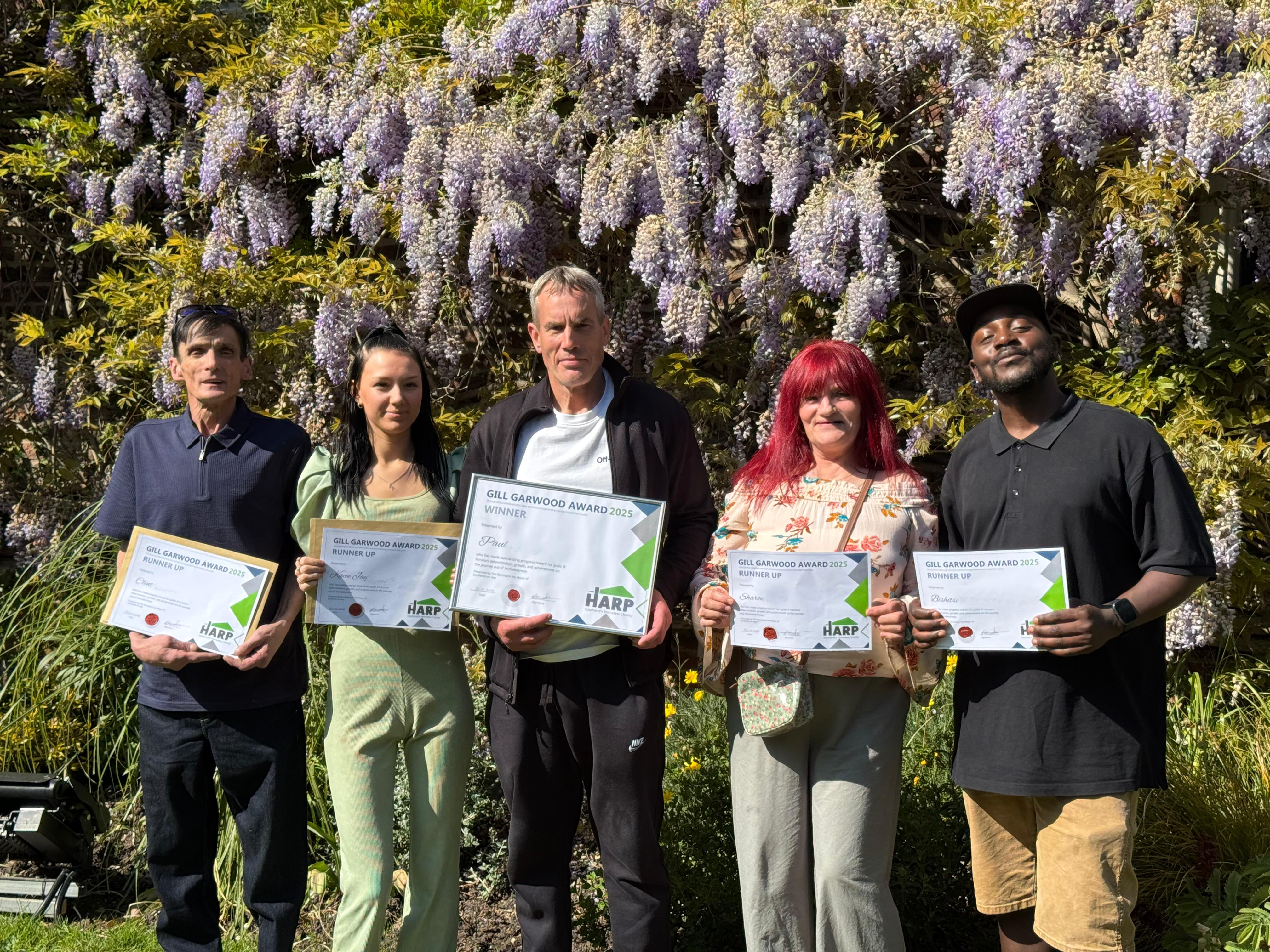 Three men and two women stand in a sunny garden proudly holding certificates