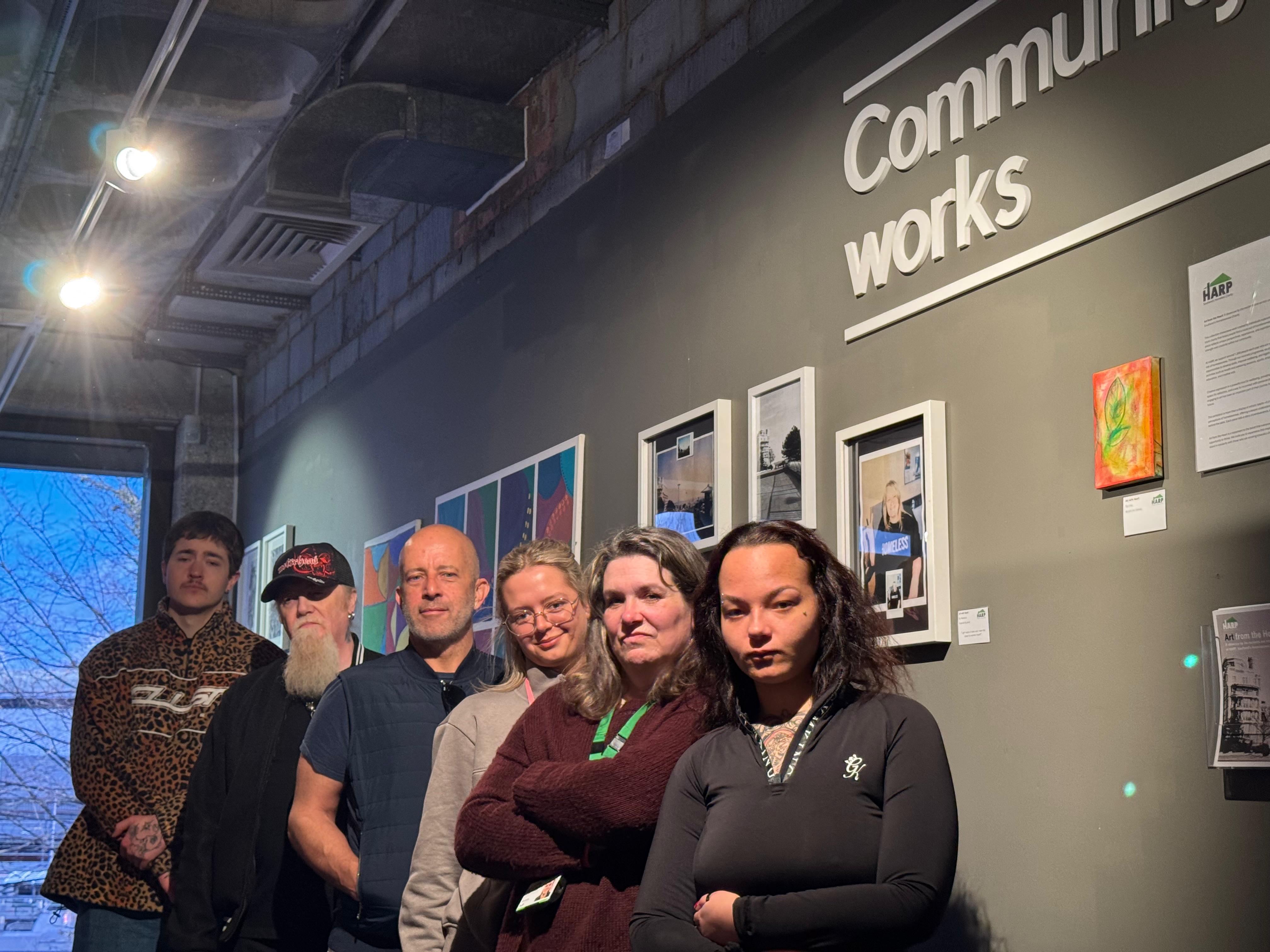 6 people stand, looking very serious, in front of art on a gallery wall.
