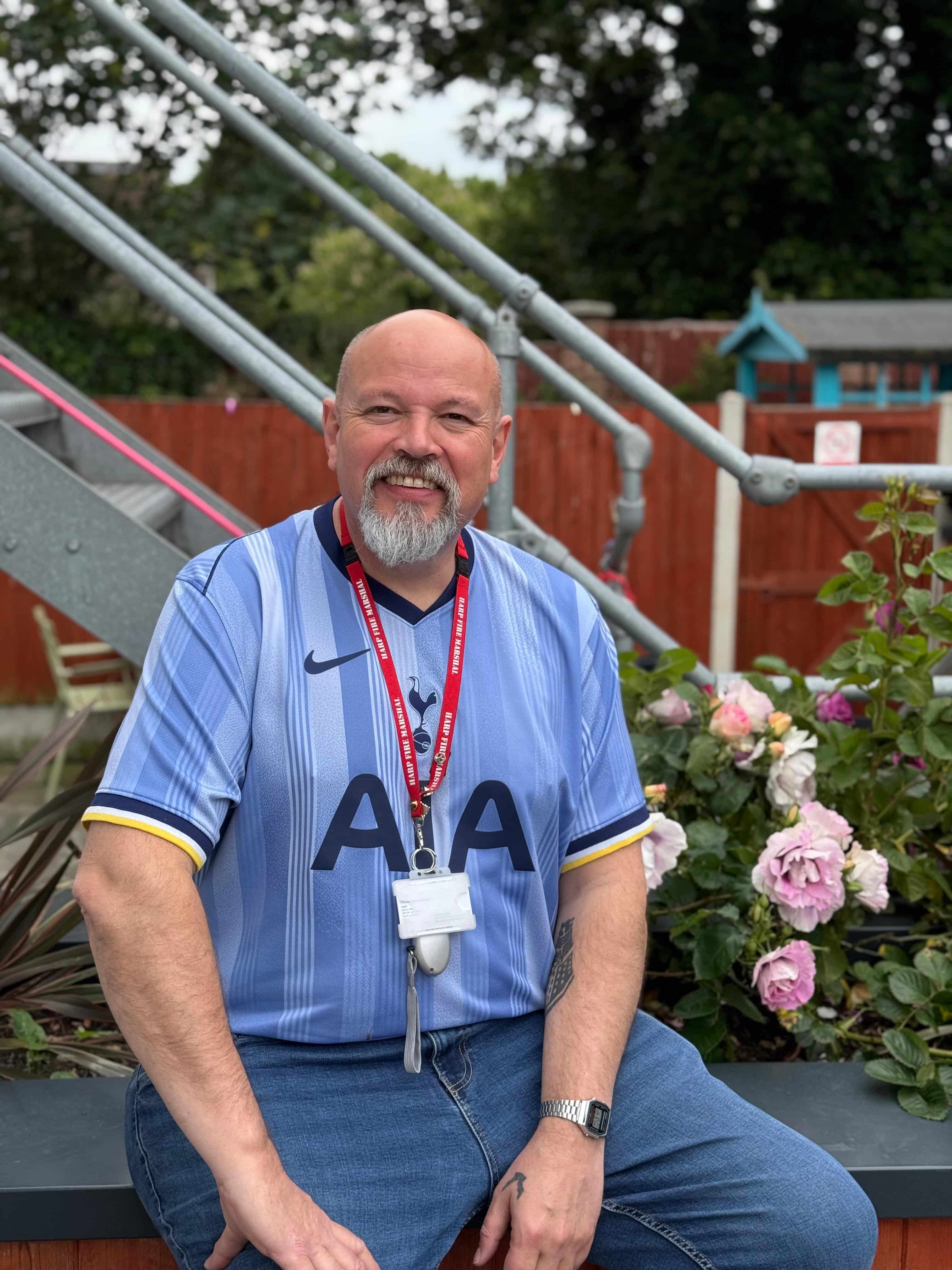 A man with a beard sitting outside. A garden can be seen in the background